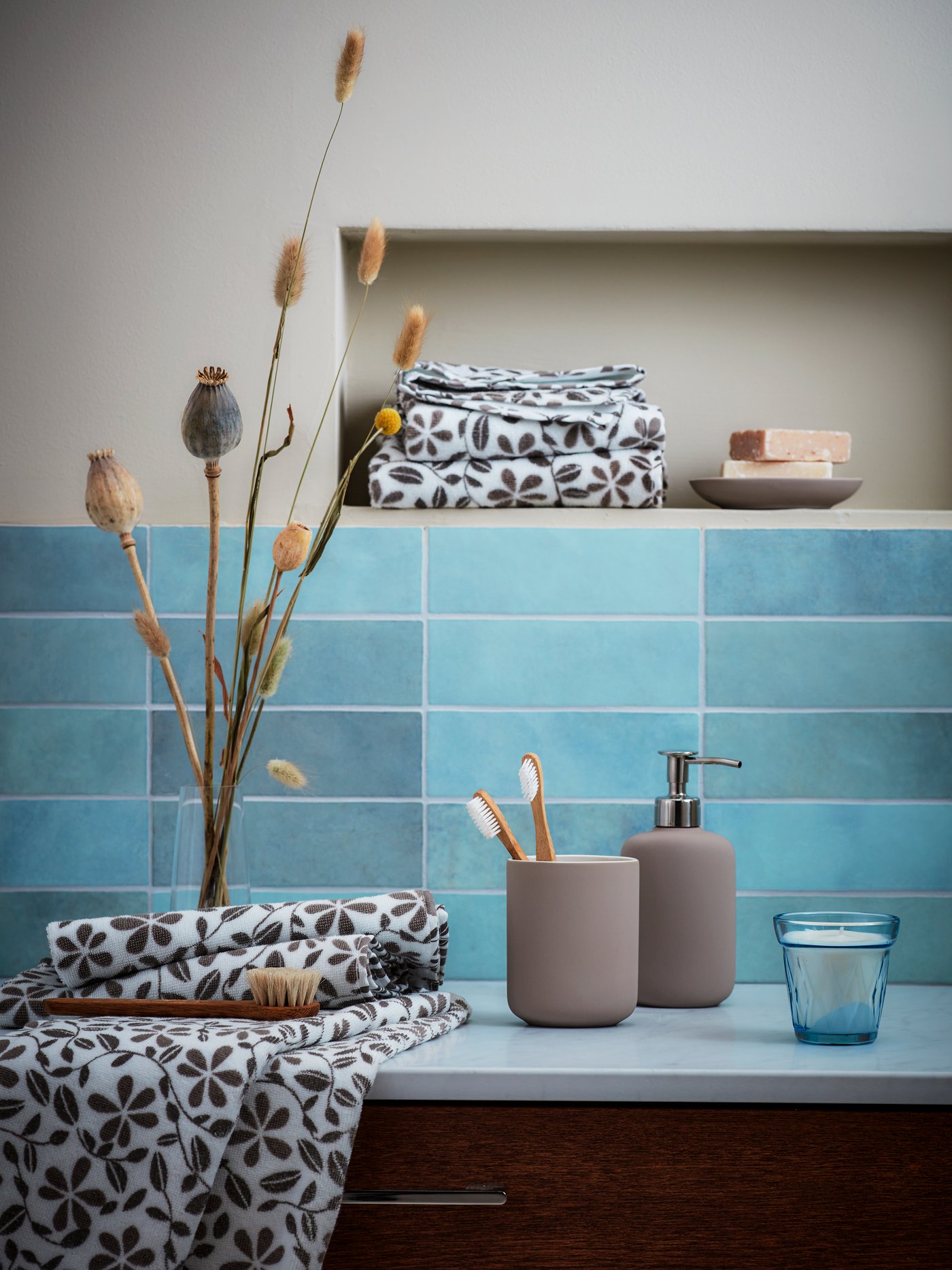 Blue-tiled bathroom section with EKOLN soap dispenser and toothbrush holder surrounded by JUVELBLOMMA towels and decorations.