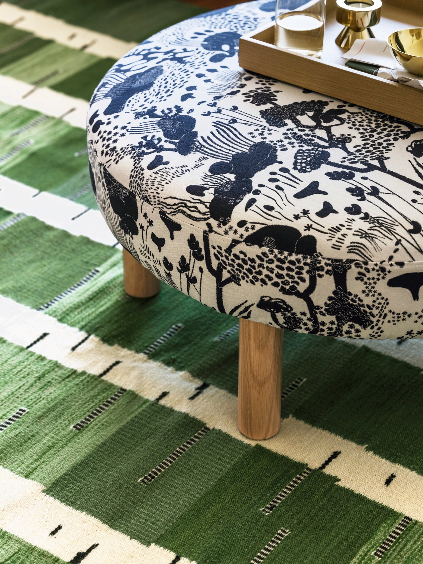 A black and white STOCKHOLM 2025 pouffe with an oak tray on top, placed on a patterned green, black and white flatwoven rug.