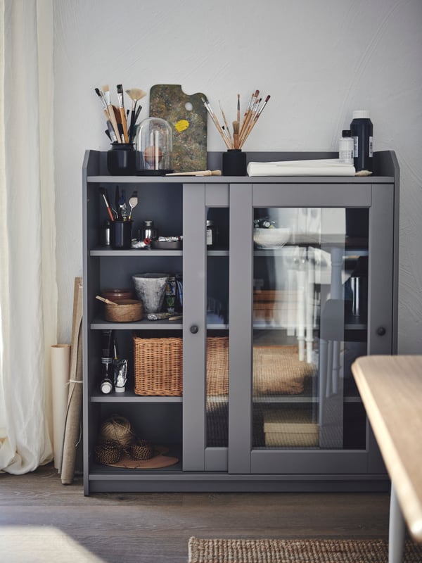 A light space with art accessories and decorative objects placed on top of and inside a grey HAUGA glass-door cabinet.