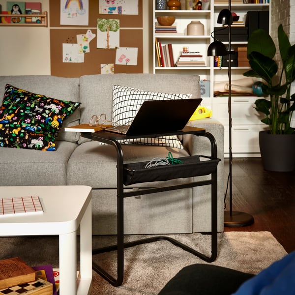 Es ist ein Wohnzimmer mit einem OLSERÖD Beistelltisch mit Laptop neben einem grauen Sofa mit gemusterten Kissen zu sehen.
