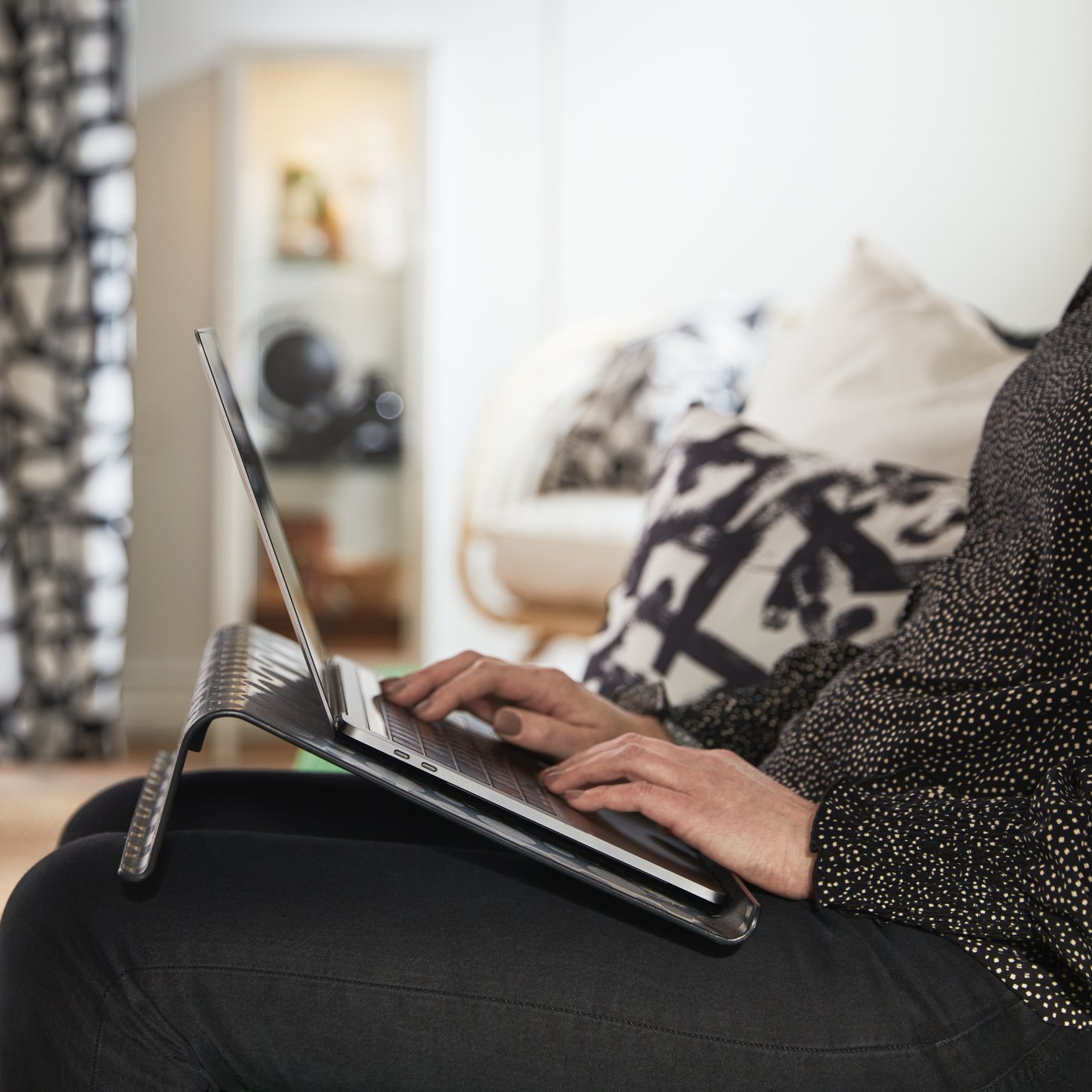 A person sitting on a sofa in a living room and using a laptop which is on a black BRÄDA laptop support.