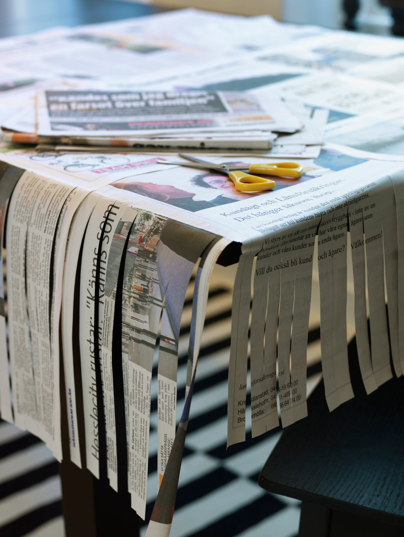 Tavolo coperto da giornali, alcuni tagliati a strisce. Forbici gialle sopra. Pavimento a scacchi. "Hasslarp" su giornale.