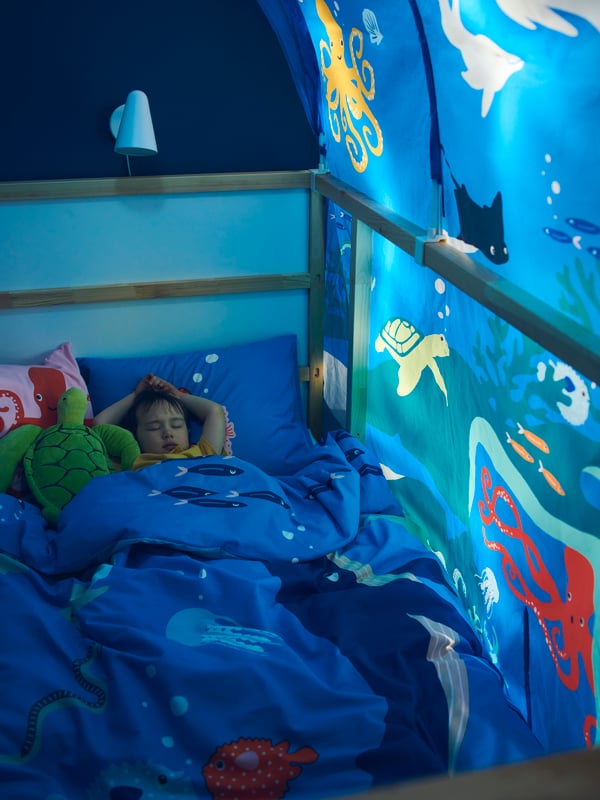 A small boy sleeps in a BLÅVINGAD ocean animals duvet cover in a KURA bed covered by a KURA bed tent.