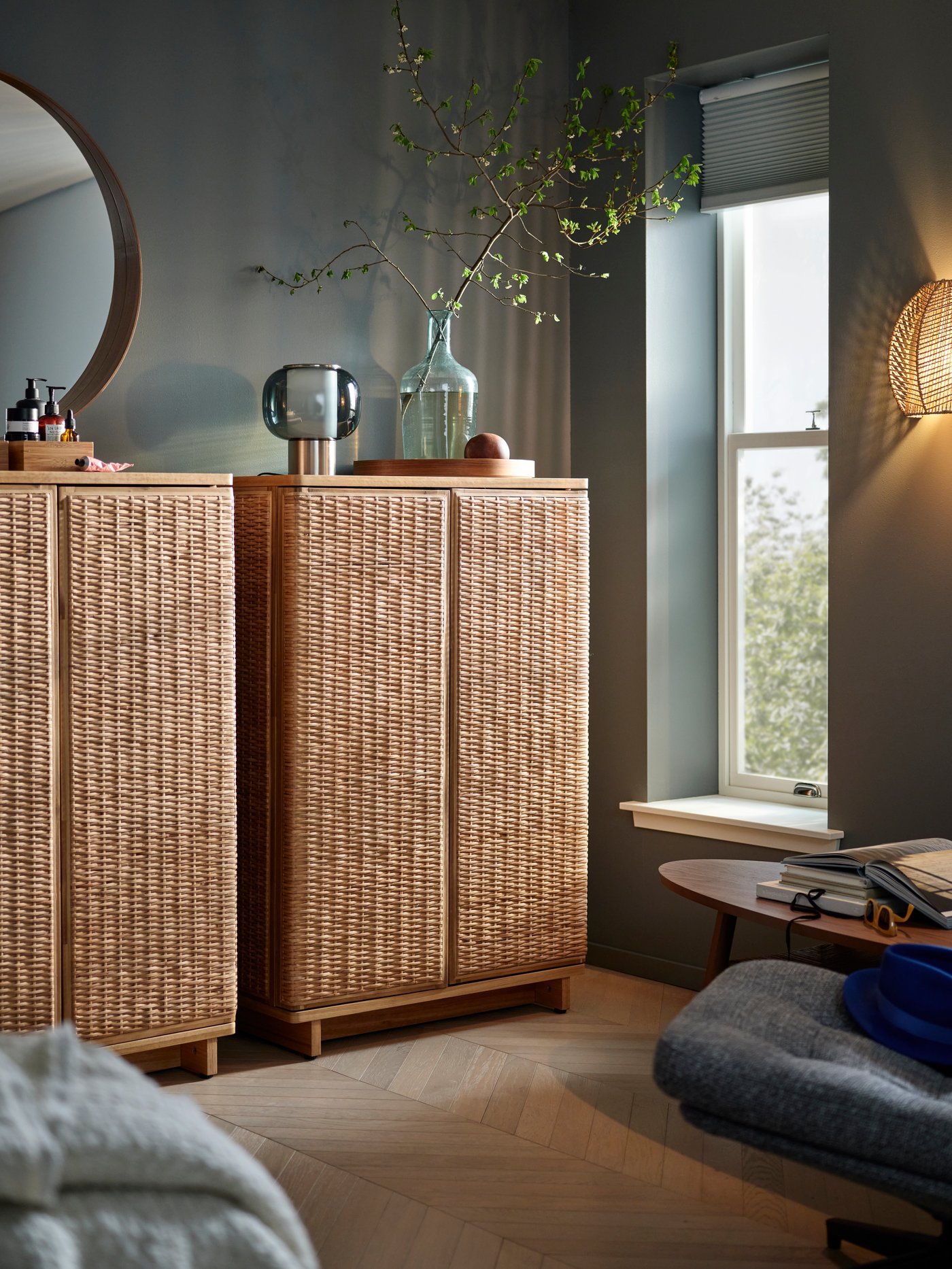 Two FRYKSÅS cabinets in rattan in a room with grey walls and wooden floor, a glimpse of a bed and an armchair in foreground.