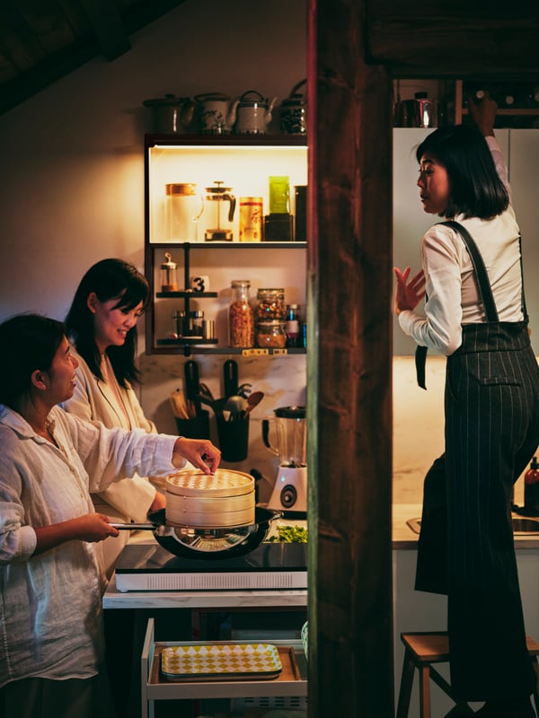 Three people in a kitchen with one person steaming food over an IKEA 365+ wok on a TILLDREDA portable induction cooktop.