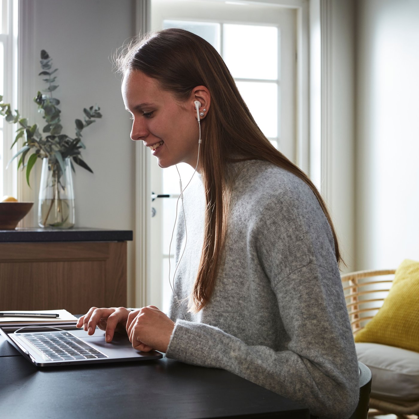 Eine Person mit langen Haaren und einem grauen Pullover sitzt an einem Tisch und arbeitet an einem Laptop. Die Person hat Kopfhörer auf.