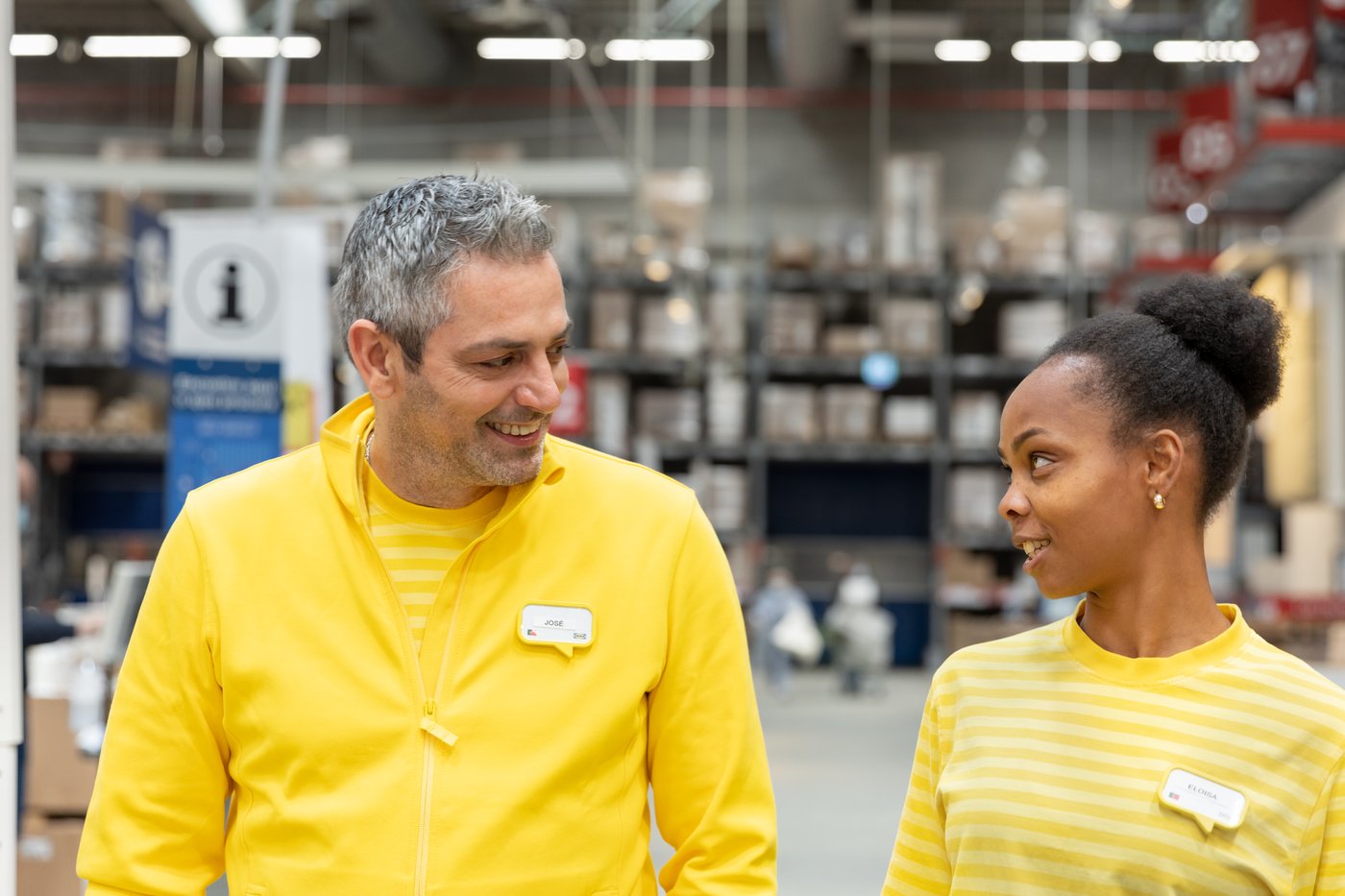 Ein IKEA Mitarbeiter und eine IKEA Mitarbeiterin gehen durch das IKEA Lager und lachen sich an.
