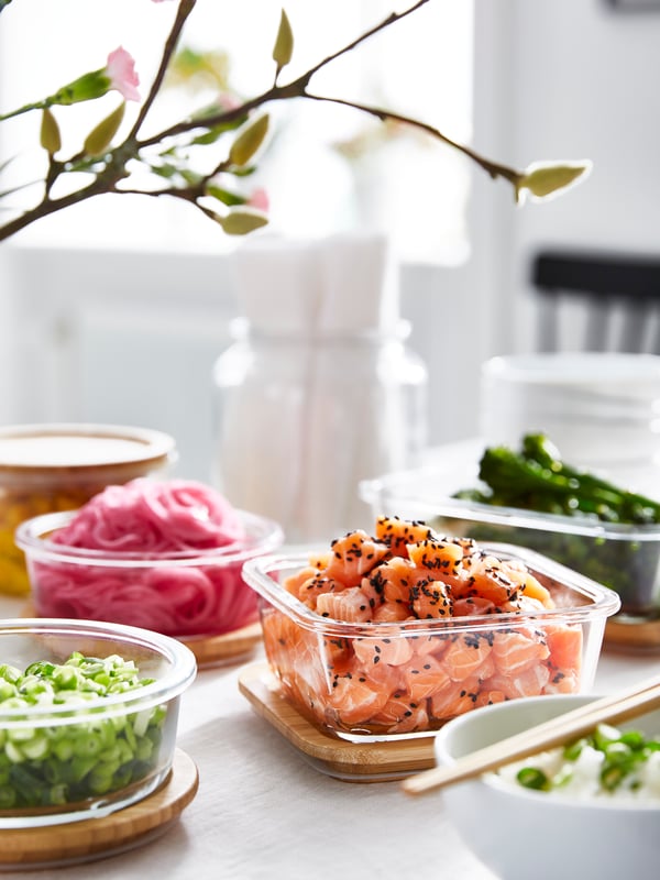Quatre contenants alimentaires IKEA 365+ en verre sont remplis de divers aliments préparés. Ils sont posés sur leurs couvercles en bambou derrière un bol de riz.
