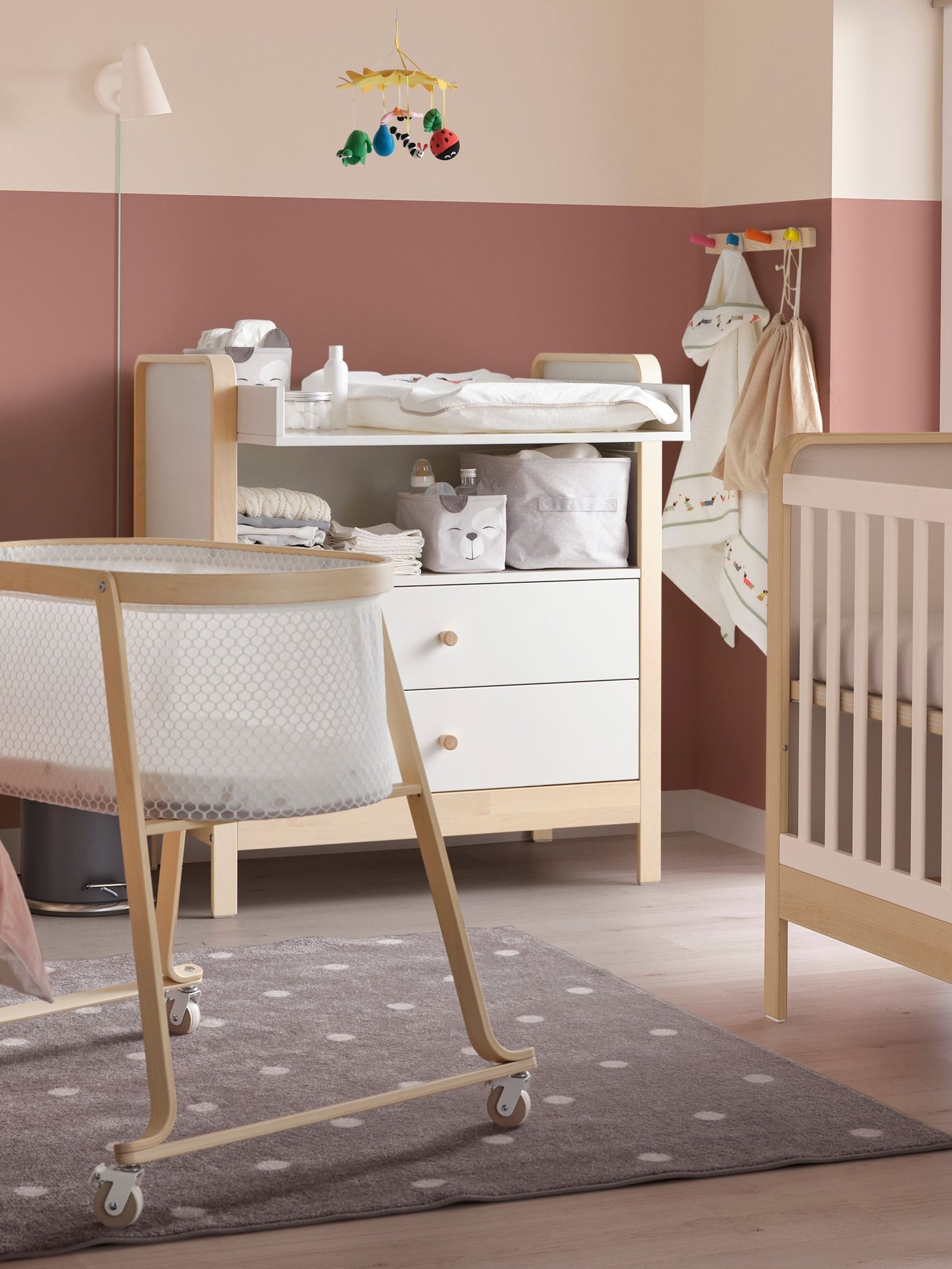 A calm nursery with light wood furniture, including a bassinet on wheels, a changing table, and a crib, on a gray polka-dot rug.