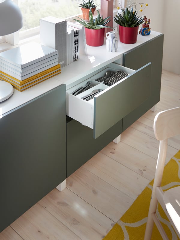 A white/grey-green storage combination with plants, books and a table lamp on its top surface. Cutlery is stored in a drawer.