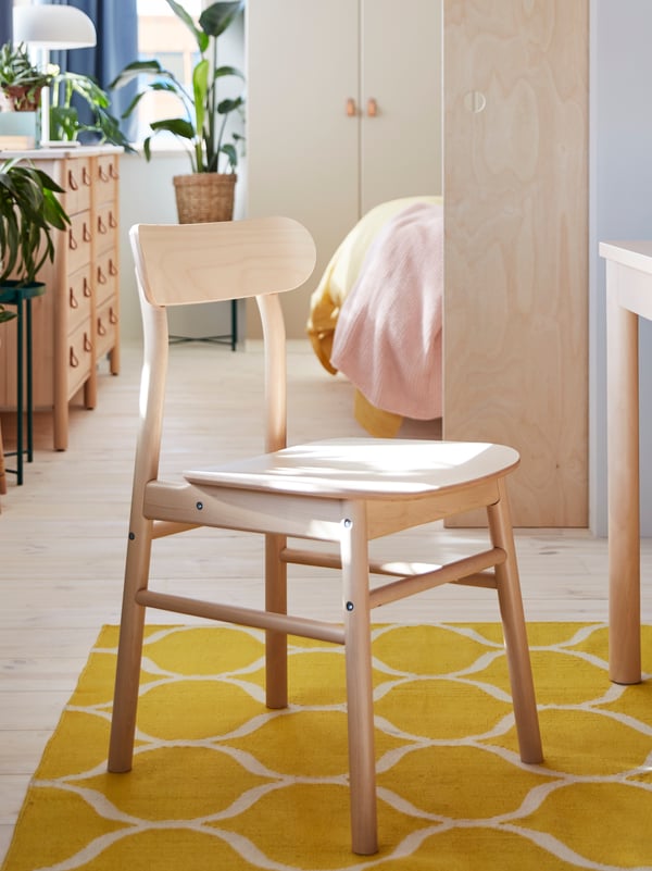 A chair in birch stands on a yellow rug. A chest of drawers in birch and green plants stand in a room in the background.