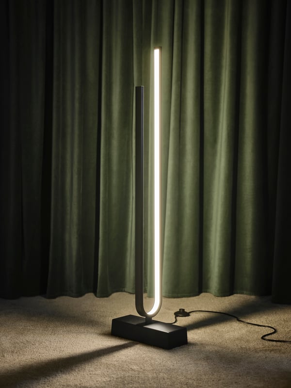 A PILSKOTT LED floor lamp that’s switched on in front of a pair of drawn grey-green SANELA room darkening curtains.