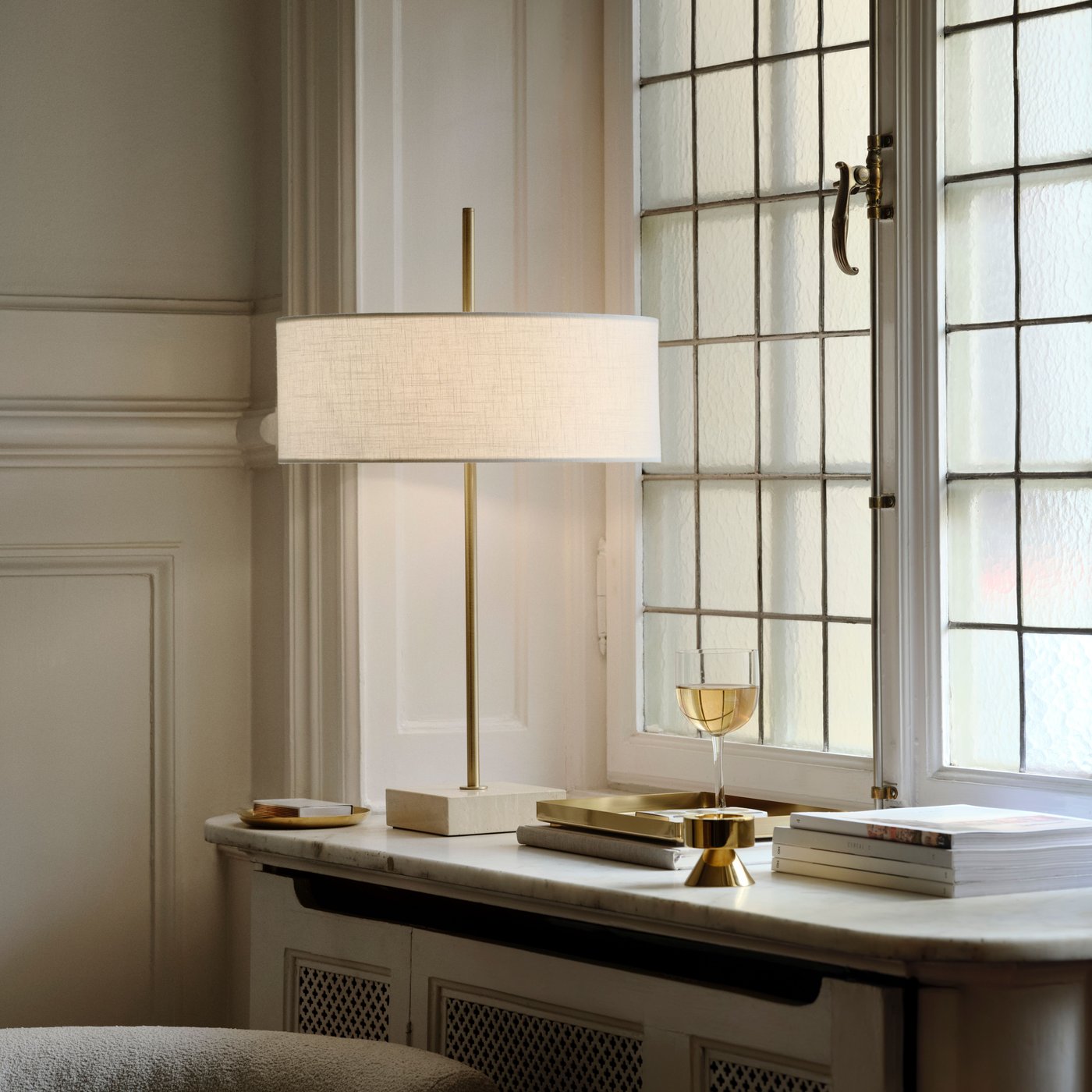 Elegant interiör med en smal bordslampa på en fönsterbräda bredvid stora spröjsade fönster. Bredvid lampan står en glasdetalj och några små föremål i mjukt dagsljus.