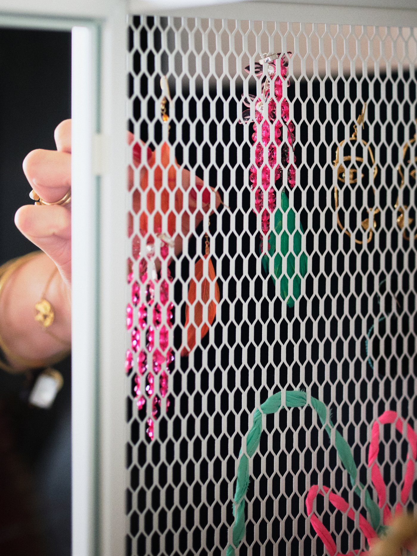 A storage unit with a vertical, white metal mesh surface. A woman’s hand is hanging several earrings in the mesh holes.