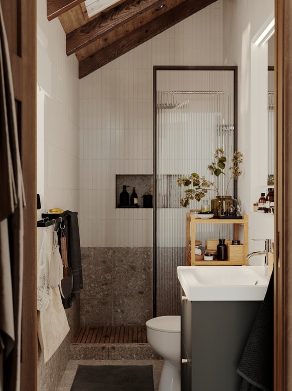 A small bathroom with white tiles, a grey stone floor and a wooden ceiling has a shower, a HAVBÄCK wash-stand and a mirror.