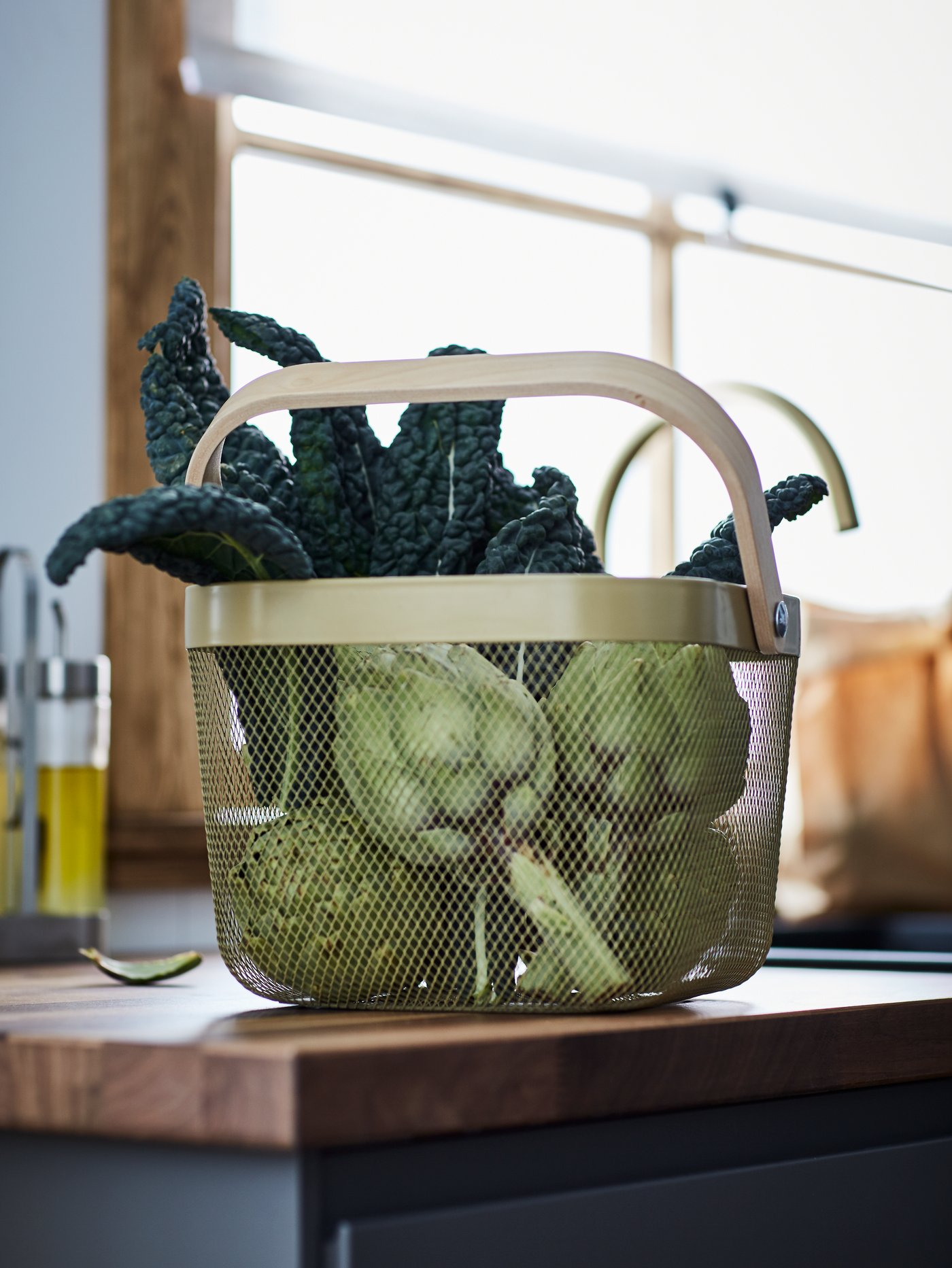A light olive-green RISATORP basket filled with black kale and artichokes, standing on a wooden kitchen top.