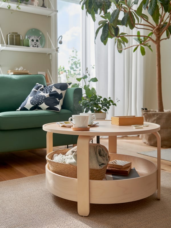 Ruang tamu yang cerah yang mempunyai meja kopi kayu birch BORGEBY, pokok berdaun lebat yang besar, sofa PÄRUP berwarna hijau gelap dan serlahan berwarna hijau yang lain.