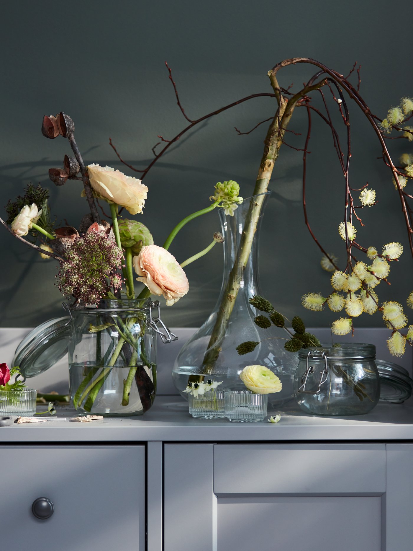 Various vase-and-flower combinations, including a single branch in a STORSINT carafe, on top of a grey HAUGA sideboard.