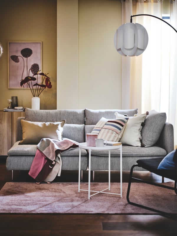 Modernt vardagsrum med grå soffa, färgglada kuddar, rosa pläd, rosa KNARDRUP matta, små vita bord med koppar och dekorativ sidobord med vas och tavlor. Ljus och inbjudande atmosfär