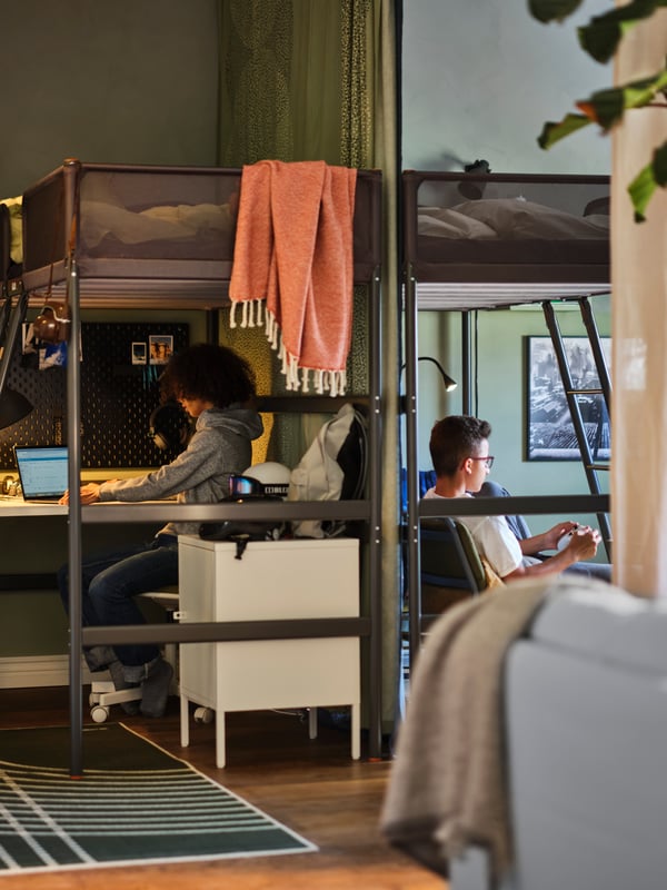 Two teenagers on either side of a sleep area, with two dark grey TUFFING loft bed frames separated by a green fabric curtain.