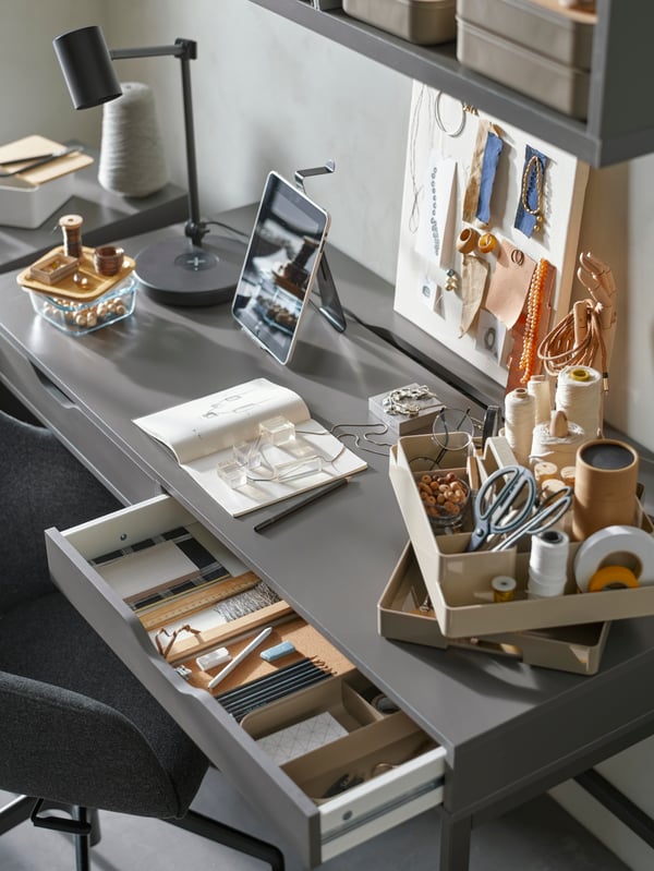 A desk with a mood board, sketchbook, supplies for making accessories, pens, and a grey-beige KUGGIS desk organiser set.