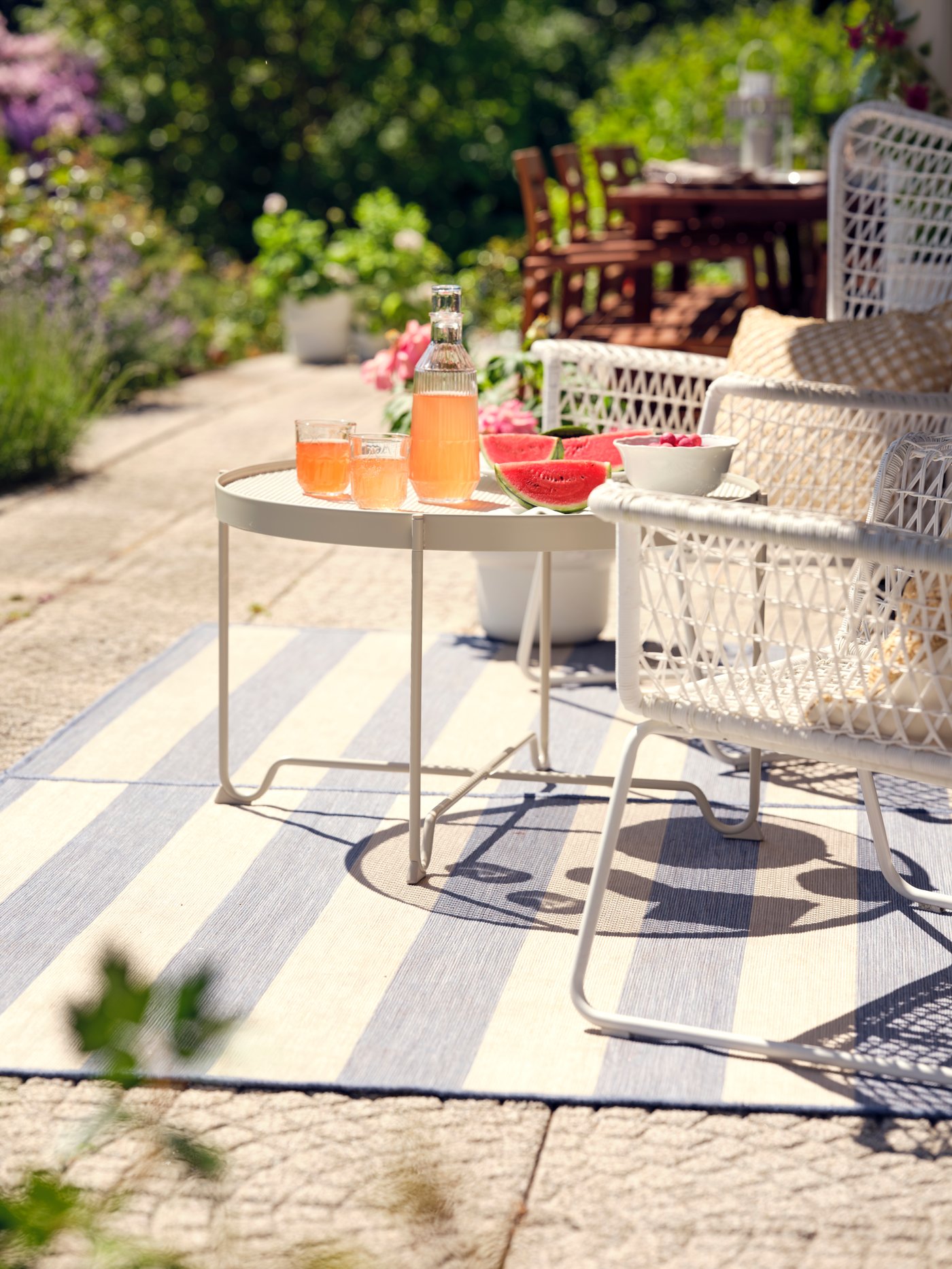 Dos sillones HÖGSTEN blancos y una mesa redonda con bebidas y rodajas de sandía encima de una alfombra VRENSTED beige/azul claro.
