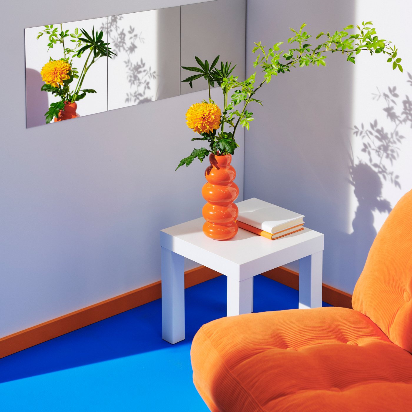 Oranžové otočné křeslo DYVLINGE z kolekce Kelinge na modré podlaze s bílým stolkem a vázou s květinou. Květiny se odrážejí v zrcadle. 