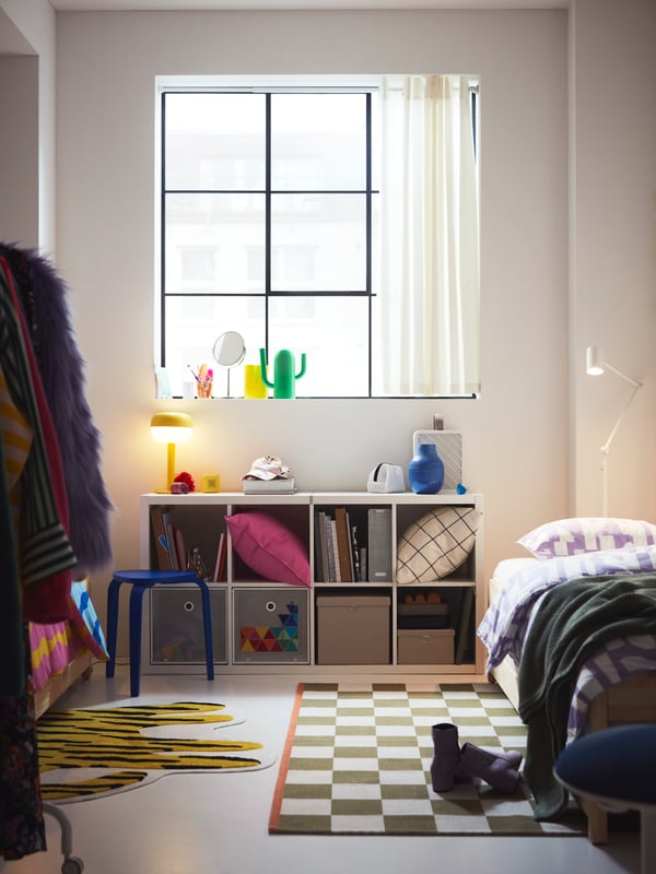 Un dormitorio infantil con un tapete IKEA y debajo de la venta un estante KALLAX con cojines y artículos en sus repisas. 