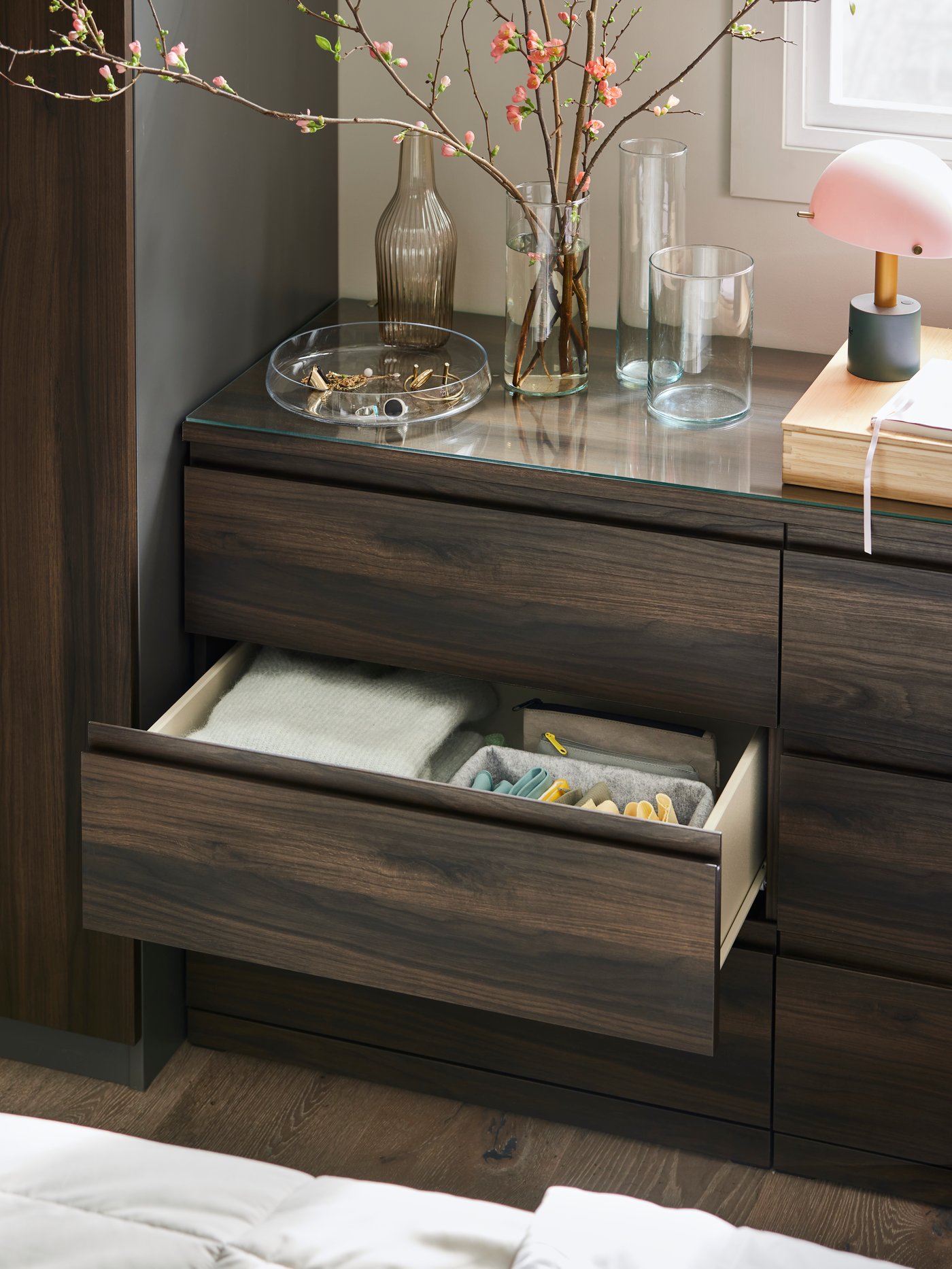 A dark brown oak effect STORKLINTA chest with 6 drawers with the middle drawer open and a lamp and vases on the top.