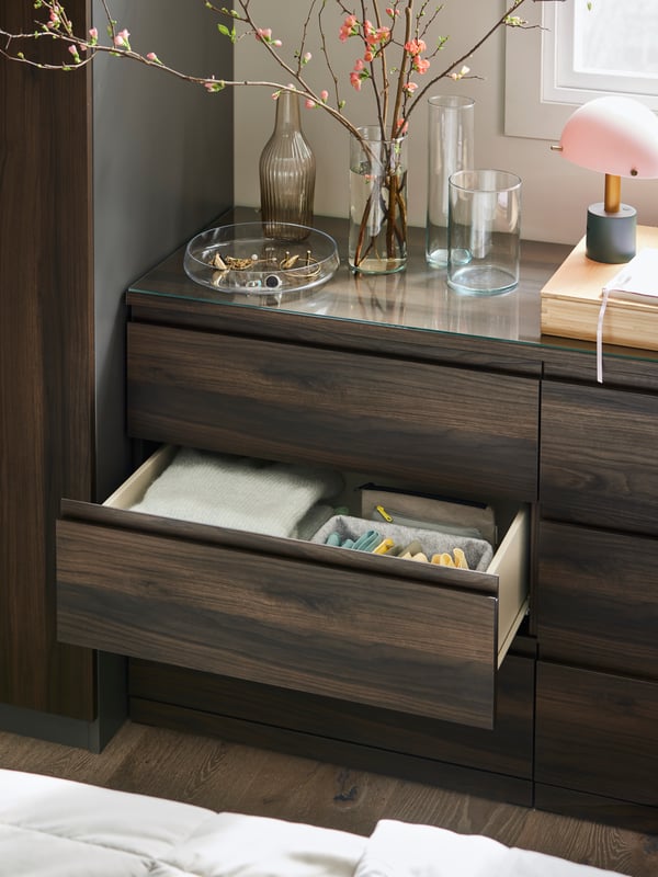 A dark brown oak effect STORKLINTA chest with 6 drawers with the middle drawer open and a lamp and vases on the top.