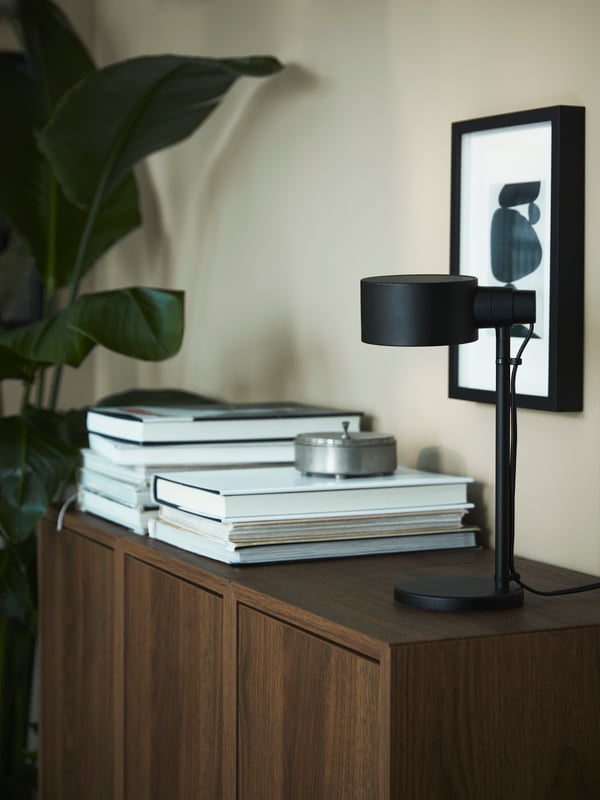 Lampe de travail LÖVMÅNAD noire et deux piles de livres sur un buffet EKET brun à effet de noyer.