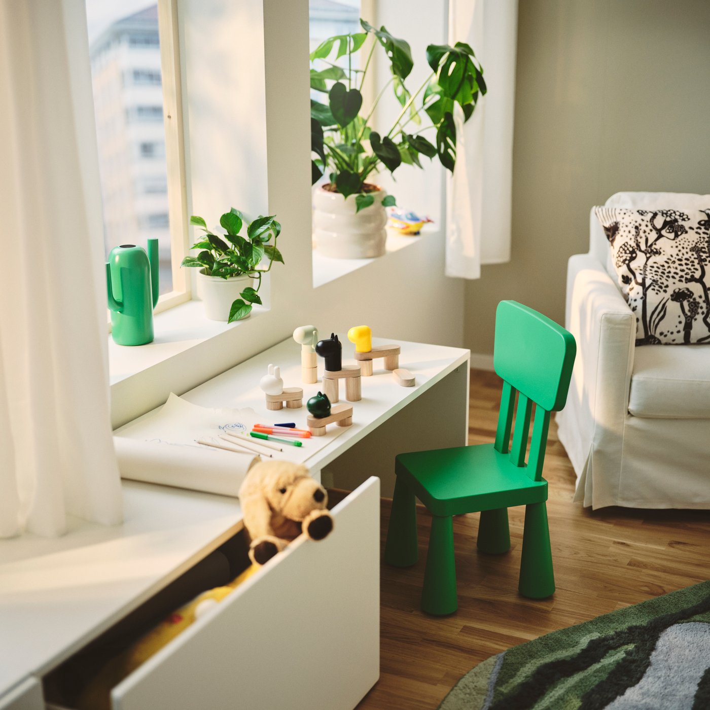 A living room featuring a white SMÅSTAD bench, surrounded by plants and a green chair, with toys on a table.