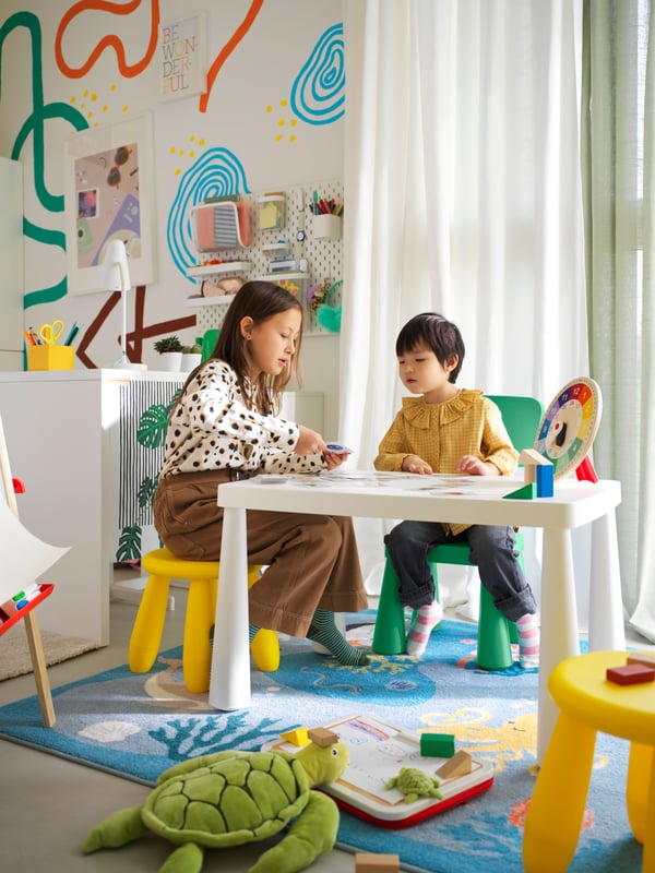 A child sits on a green MAMMUT children’s chair at a table with an adult who is sitting on a yellow MAMMUT children’s stool.