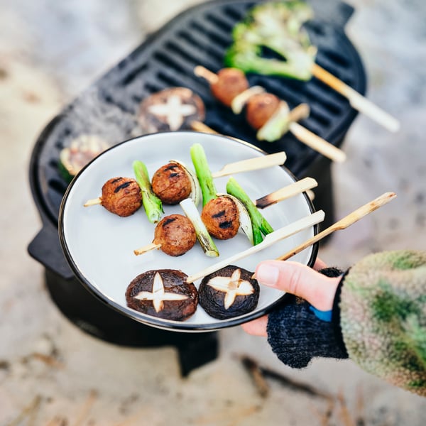 A hand holds up a SOLUPPGÅNG plate with mushroom and plantball skewers on it, close to a SOLUPPGÅNG barbecue.
