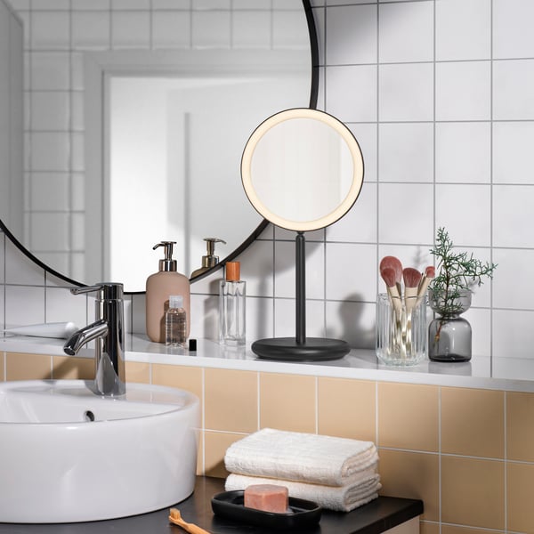 The magnifying side of a black, dimmable EKFÄNN table mirror with integrated lighting is lit, on a bathroom ledge by a sink.
