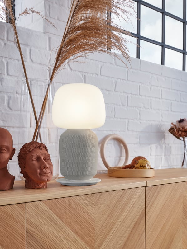 Buffet en bois clair avec une lampe de table grise et opaline et un grand vase en verre transparent devant un mur en briques blanches