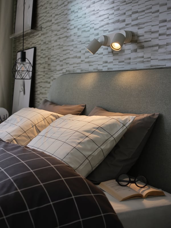 White/black check quilt cover and pillowcases, a white double wall lamp, an open book and black reading glasses.