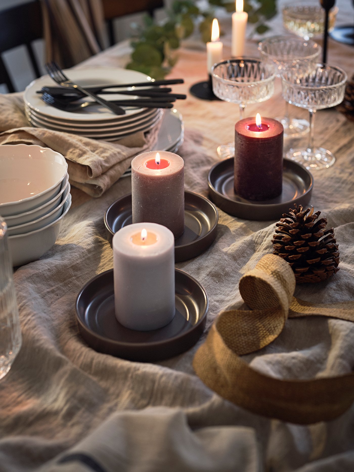 A dining table with sets of dinnerware and cutlery, and scented top candles placed on flat candle trays.