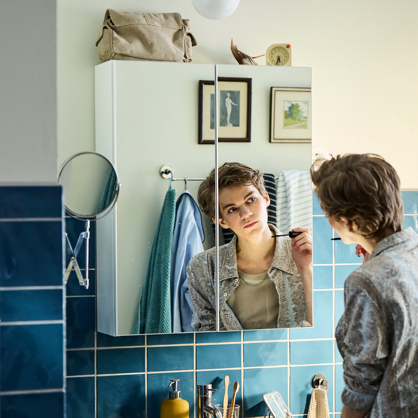Eine Person in einem blau gefliesten Badezimmer betrachtet ihr Spiegelbild in einem ENHET Spiegelschrank mit 2 Türen.
