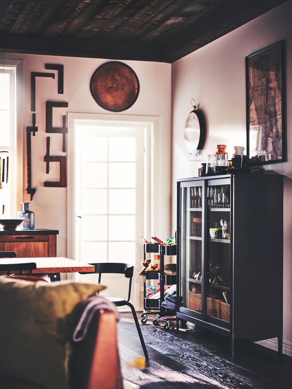 Un área de comedor con una silla YNGVAR, una vitrina IDÅSEN gris oscuro con puertas corredizas y dos carritos RÅSKOG negros.