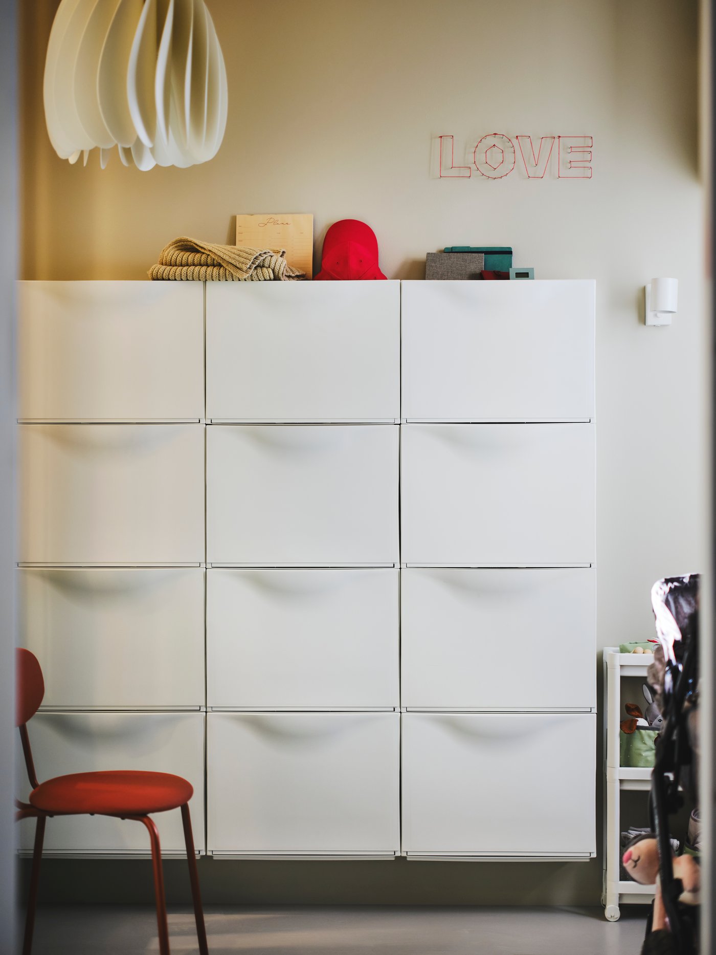 There are six white TRONES storage units on a wall, with a red-brown ÖSTANÖ chair in front of them. A scarf and a hat lie on top of the storage units.