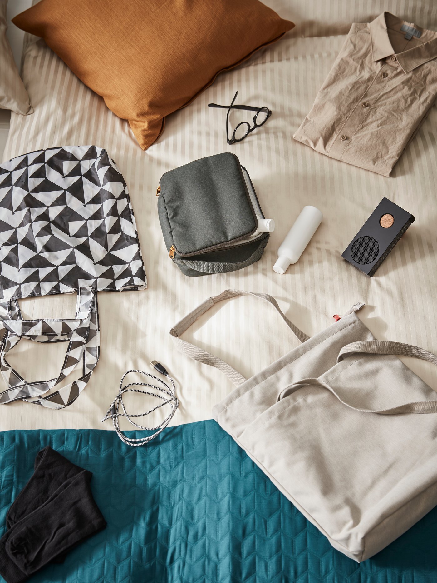DRÖMSÄCK travel bags lying on a bed next to a tote bag, a USB cable, a pair of glasses, a shirt and a portable speaker.