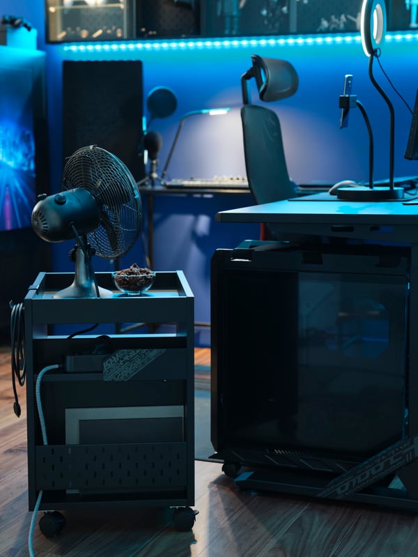 An UPPSPEL CPU stand and drawer unit by a desk in a blue-lit gaming room. There’s a fan and snacks on the drawer unit.
