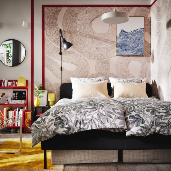 A dark grey SLATTUM upholstered bed in the corner of a bedroom with two duvets covered in BROKDRACENA bed linen on it.