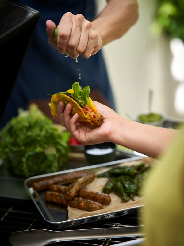 한 사람은 속을 채운 타코 쉘을 들고 있고 다른 사람은 즙을 짜고 있는 두 사람, 그리고 GRILLTIDER 그릴티데르 바비큐 도구와 KORVMOJ 코르브모이 베지 핫도그가 있는 이미지.