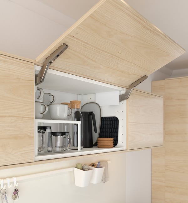 Un mueble mural de cocina METOD con la puerta abierta hacia el cielo. Dentro del mueble hay tazas, una tetera y otros recipientes de vidrio.