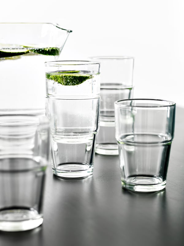 Several REKO clear glasses, some are stacked and sparkling water is being poured into one glass with a slice of lime inside.