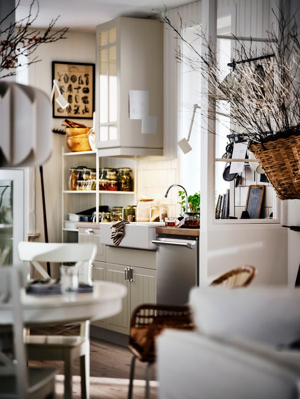 Une cuisine et une salle à manger beiges et lumineuses de style rustique avec des armoires blanches, une table ronde et des branches séchées décoratives.