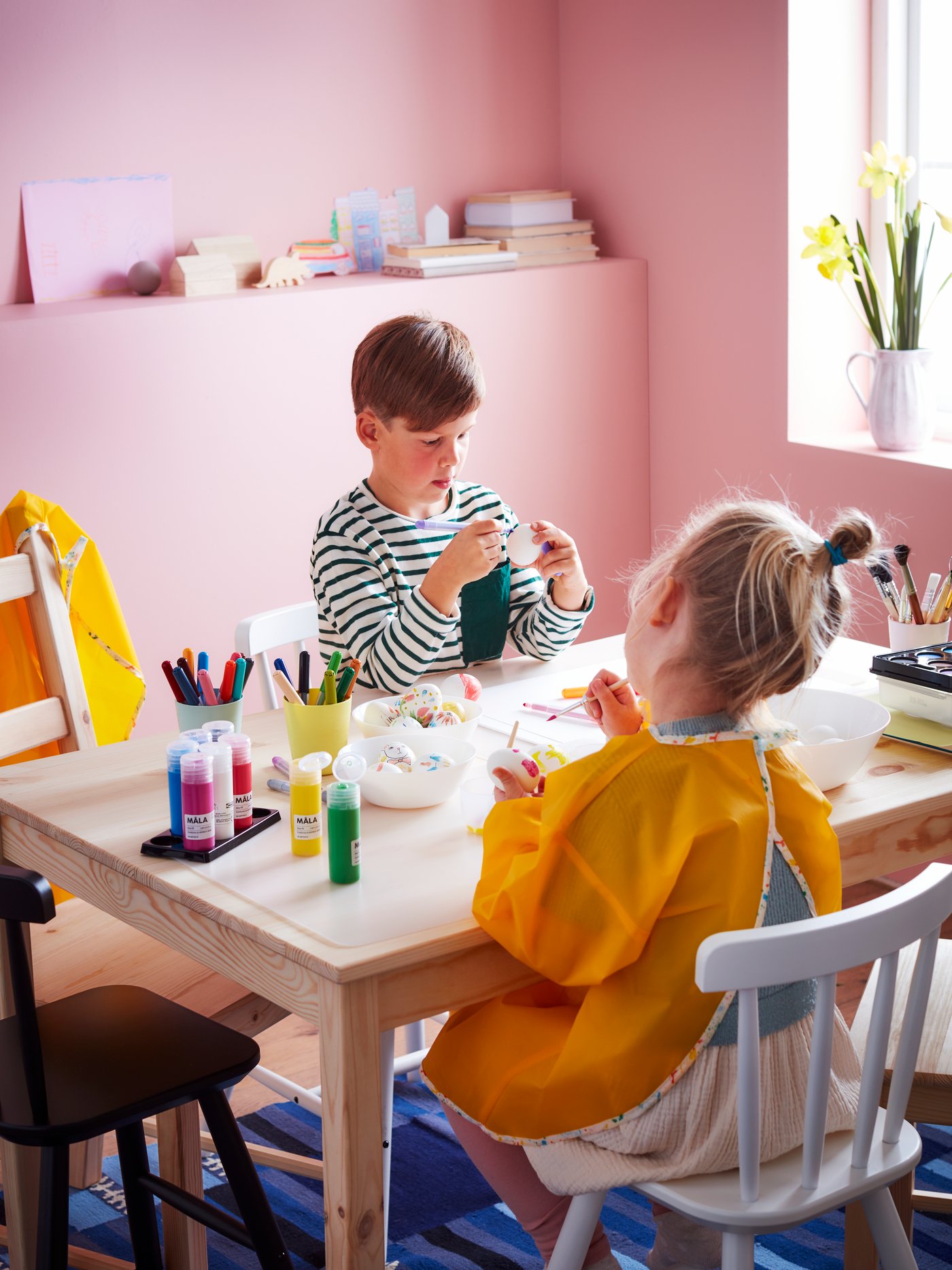 Two children sit at a low table, engrossed in colouring eggs. The table is filled with eggs and MÅLA felt-tip pens and paint.