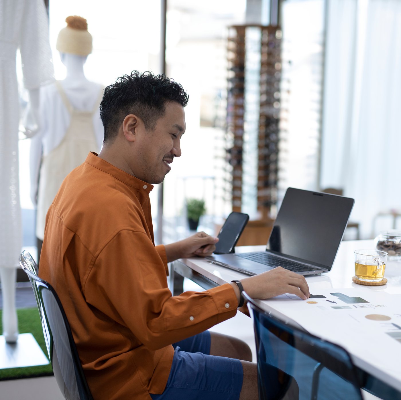 A person sits in front of a laptop on a desk while holding a smartphone.